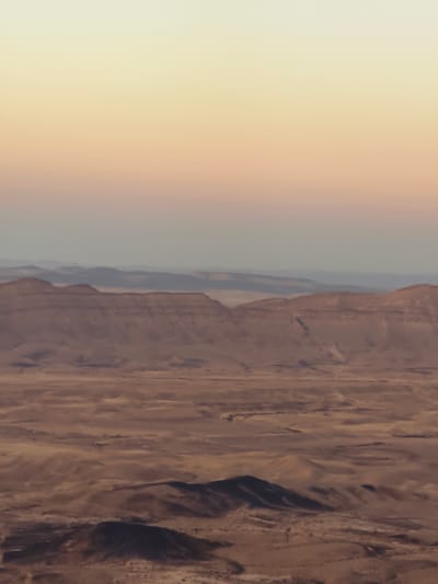The image depicts a desert landscape with multiple layers of hills and mountains receding into the distance. The sky above transitions from a pale yellow at the horizon to a soft, peachy orange higher up, suggesting a sunset or sunrise. The terrain is arid and barren, characterized by shades of brown and tan, with subtle variations in texture and topography. In the foreground, darker, more pronounced mounds of earth are visible, some casting shadows. The overall scene conveys a sense of vastness and natural beauty. This is the Ramon Crater in Yeruham, Israel.