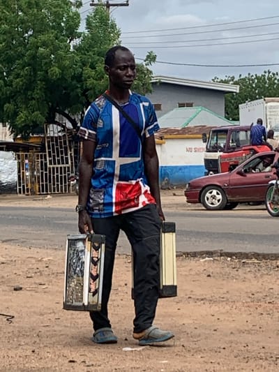 This image captures a street scene in Shendam, Nigeria, featuring a dark-skinned man standing on a dirt shoulder to the right of a paved road. The man, with short dark hair and a neutral expression, wears a blue, white, and red short-sleeved shirt with a prominent cross-like design, dark trousers, and grey-blue open-toed sandals. A dark strap crosses his left shoulder, and a dark watch is on his left wrist. He carries two identical rectangular, box-like containers, one in each hand, at waist level. These containers appear to have transparent panels revealing contents and feature textured or patterned sides; the container in his right hand displays black arrow-like patterns.
The ground beneath him is dry, light brown earth mixed with small stones and debris, transitioning to a darker paved road. In the middle ground, a maroon sedan with a five-spoke white alloy wheel is visible on the road, facing left. Behind it, a red and white truck is partially obscured, with an individual in a blue plaid shirt seated on its rear. Another person is partially visible to the right of the truck. On the far right, a bicycle or motorcycle wheel with green and red accents is visible.
The background on the left features dense green trees and a light-colored metal gate structure. Buildings with corrugated roofs and painted walls are visible throughout the background. A white wall with partially visible dark text "NO SMOKING ON" is present behind the sedan. Overhead, multiple power lines stretch horizontally across a light grey, overcast sky.