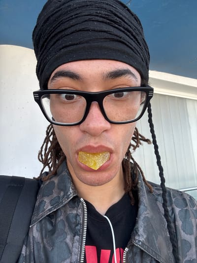 A person wearing glasses and a black head covering has a triangle-shaped yellow gummy candy in their mouth. The person is also wearing a black and gray patterned jacket over a black shirt. Their hair is styled in dreadlocks. The background appears to be an outdoor setting with a white wall. There are no indications of the location being in San Bartolomé, Spain.