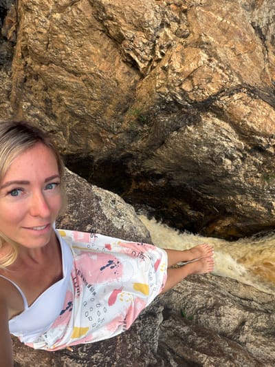 A woman with blonde hair and blue eyes is taking a selfie outdoors. She is wearing a white bikini top and a patterned sarong. Her feet are visible, resting on a rock near a flowing body of water. The background consists of large, textured rock formations. The scene appears to be a natural environment, possibly a waterfall or river.