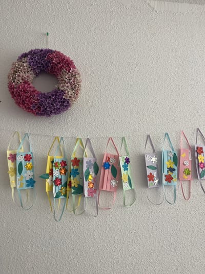 In a well-lit indoor setting, likely a school or community center in Geneva, Switzerland, a white textured wall serves as a backdrop for a decorative display. The focus is on a vertical arrangement of seven colorful, handmade face masks, each adorned with various paper flowers and green leaf cutouts. These masks appear to be a craft project, possibly created by children, as evidenced by the playful and varied decorations. Below the masks, to the left, hangs a circular wreath composed of numerous small, artificial flowers in shades of pink and purple. A tiny green tack is visible on the wall near the wreath. The masks themselves are each attached to the wall by thin, colorful ribbon loops, suggesting they are either for display or part of an activity. Some masks have small handwritten labels with text in Cyrillic script, indicating potential origin or names associated with the creations. The overall atmosphere is cheerful and crafty, with a sense of community or educational engagement. The lighting suggests daytime indoors, with no discernible weather cues.
