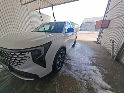 A white SUV is being washed at an outdoor car wash located in Shamkhal, Russia. The car is positioned under a corrugated metal roof, with its front end angled towards the viewer. Water and foam are visible on the ground around the vehicle, indicating the washing process is underway. The car has its windshield wipers raised. In the background, a two-story building with multiple windows and a metal roof is visible, along with a small truck parked to the left of the building. A metal fence encloses the car wash area. The sky is clear and bright.