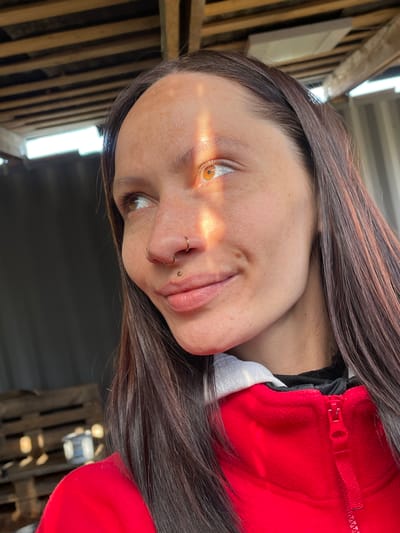 A woman with brown hair and amber eyes, wearing a red jacket over a white and black top, is photographed. She has piercings in her nose and lip. Light streams across her face. The setting appears to be outdoors, near wooden beams and a corrugated metal structure, possibly a shed or shelter. The precise location in Doma 45 Kilometr, Russia, cannot be determined from this image alone.