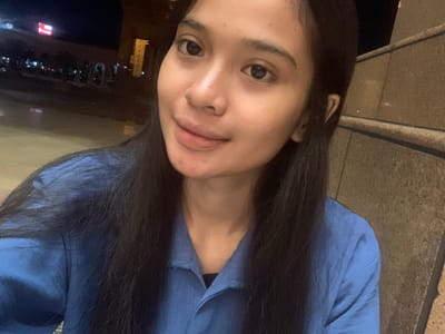 The image is a close-up selfie of a young woman, captured in a low-light setting. The photograph is oriented vertically, showing the woman's head and shoulders as she looks directly at the camera with a neutral to slightly smiling expression. Her fair-skinned face is relatively well-lit, highlighting her dark eyes and long, dark hair that frames her face. She is wearing a blue collared top or blouse.

To her left, the background consists of a textured, light brown or grey surface, possibly a stone wall or ground. To her right, the scene recedes into a dark, out-of-focus area punctuated by blurred urban lights, including red and white glows, which are characteristic of an outdoor, night-time environment in a city. This suggests the photo was taken outdoors in the evening or at night. The location context provided indicates this scene is in Samarinda, Indonesia. No discernible text is visible within the image.