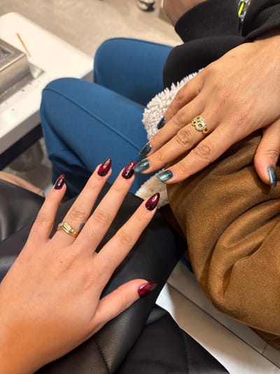 Two pairs of hands display manicured nails. One set features an emerald green cat-eye effect polish. The other showcases a striking red and black gradient design on pointed nails. Both hands are adorned with golden rings. The fingers rest on a backdrop of blue pants, a brown garment, and a black leather material. A portion of a white surface is also visible, likely a table or counter.