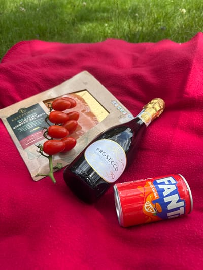 A picnic spread is laid out on a bright red blanket on a grassy area. In the image, there is a bottle of Prosecco, a can of Fanta Fruit Twist, a package of smoked salmon, and a vine of cherry tomatoes. The Prosecco bottle is dark green glass with a gold foil cap and a pale label featuring ornate text. The Fanta can is red with the Fanta logo and images of fruit. The smoked salmon package is a light brown cardboard with a clear plastic window revealing the salmon. The cherry tomatoes are bright red and still attached to a green vine. The background is a blur of green grass. The location is the town of Mid Sussex, United Kingdom.