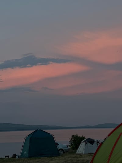 This media file shows a campsite by a body of water, likely during sunset. The sky is filled with streaks of pink and grey clouds, indicating a soft, diffused light. The scene is set in Колодезный, Russia.
The primary objects in the foreground are two tents. One is a dark blue/green tunnel tent, set up and visible from the side. The other tent is partially visible and appears to be light-colored with a dark entrance. A portion of a car is visible near the tents, its tire and wheel clearly discernible. In the background, a tree line or low hills are visible along the horizon, separating the land from the sky. A distant body of water, possibly a river or lake, is present, reflecting the muted colors of the sky. A sliver of a bright green and red tent is visible in the upper right corner, framing the scene.
The time of day is clearly evening, with the soft, pastel colors of the sky suggesting either sunrise or sunset. The weather appears calm, with no indication of wind or rain. The setting is a natural, outdoor environment, ideal for camping.
No people are visible in the media file, and no text is discernable. The overall mood is peaceful and serene, evoking the quiet atmosphere of a campsite at dusk.