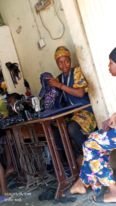 This image depicts an indoor tailoring workshop, specified as located in Gama, Nigeria, captured on 2026/03/19 at 15:01. Three individuals are visible. A young female, centrally positioned, wears a patterned headwrap and a dark blue garment over a vibrant yellow, green, and orange patterned fabric, with a yellow measuring tape draped around her neck. She is seated at a wooden sewing machine table, actively manipulating pieces of dark blue fabric and another with a purple and black pattern. To her immediate left, a partially visible individual is operating a black manual sewing machine. To her right, a third person is partially visible, wearing blue trousers adorned with a prominent floral pattern and dark slide sandals on a concrete floor. The interior features light-colored, worn walls with visible cracks, exposed white electrical wiring, and a white electrical switch. The central sewing machine table features an ornate, dark metal base with a visible foot pedal mechanism. Fabric remnants and dark bundles are present on the floor and in the background.