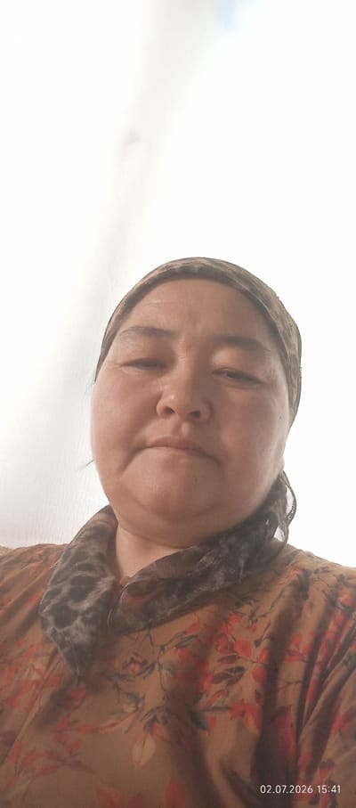 A medium shot, from a low angle, shows a woman in Chon-Kara, Kyrgyzstan. The woman is wearing a brown and orange floral print shirt and a patterned headscarf. Her face is mostly visible, and she appears to be looking slightly down and to her left. The background is a plain, off-white surface, possibly a wall or curtain. A timestamp in the lower right corner reads "02.07.2026 15:41".