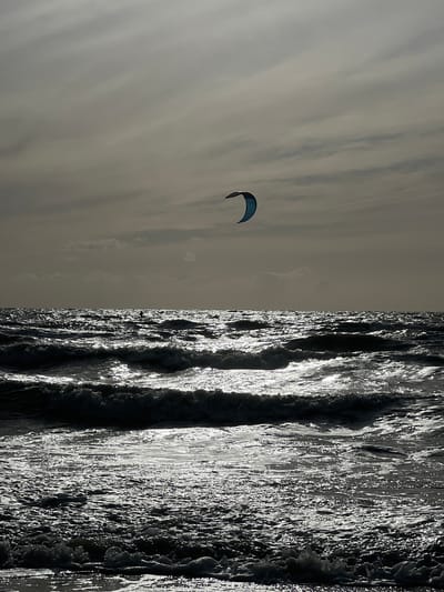 A kite surfer is captured mid-air over the choppy waves of Deauville beach, France. The scene is dramatic, with dark, turbulent water reflecting the overcast sky. The kite surfer, a dark silhouette against the bright water, is suspended in the air, their kite a bright blue crescent against the muted sky. The waves are rolling in, creating a textured surface on the water, with streaks of light glinting off the crests. The sky is filled with layers of clouds, creating a sense of depth and atmosphere.