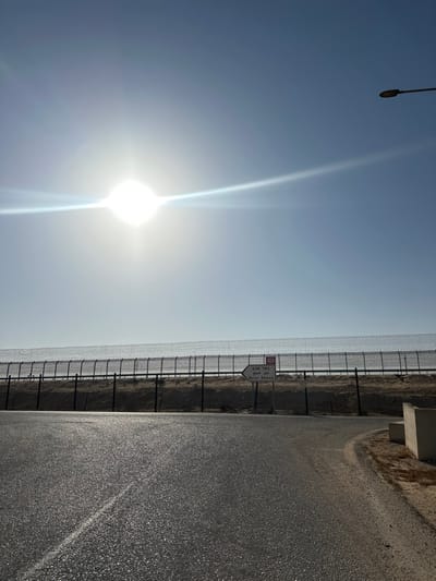 The image displays an outdoor daytime scene in the Ramat Negev Regional Council, Israel, specifically along Route 10, indicated by a directional sign for Be'er Sheva. A bright, flaring sun, positioned in the upper left of a clear, light blue sky, illuminates the scene and creates a prominent horizontal lens flare. In the foreground, a dark asphalt road surface, reflecting intense sunlight, extends from the bottom of the frame and curves slightly to the right. A dirt shoulder and a partial light-colored concrete barrier are visible along the right edge. The midground features a tall, multi-layered security fence, comprising a dark metal framework topped with dense rolls of barbed or concertina wire, spanning horizontally across the frame. Behind this fence, a light brown, elevated earth embankment is visible. A white directional sign, positioned in front of the fence slightly right of center, displays a black left-pointing arrow and text in Hebrew ('באר שבע'), Arabic ('بئر السبع'), and English ('B'er Sheva'), accompanied by a red square '10' route marker. A dark street light fixture extends into the upper right corner of the frame. No people or vehicles are present.