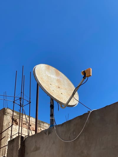 A low-angle shot depicts a satellite dish mounted on the exterior of a building. The dish is circular and off-white with visible weathering. A feedhorn in a tan housing is positioned at the dish's focal point. The dish is attached to the building with metal supports. Below the dish and mounting, exposed rebar protrudes from the building's concrete structure. A coaxial cable runs from the dish to the building. The sky is a clear, bright blue. This image was taken in the city of Sidi Chahmi, Algeria.