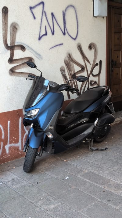 The image shows a blue Yamaha NMAX scooter parked on a cobblestone sidewalk next to a cream-colored wall covered in graffiti. The scooter is the primary subject, angled towards the camera. It appears to be in good condition. The graffiti on the wall includes purple and brown spray-painted words and symbols, adding a gritty urban element to the scene. The scooter is positioned in front of a section of the wall that is painted with a reddish-brown color at the bottom. Above the scooter, a wooden-paneled overhang or door is visible. The sidewalk is made of gray cobblestones, and a few pieces of litter, possibly cigarette butts, are scattered on the ground near the scooter's rear wheel. The lighting suggests it is daytime, with shadows visible on the cobblestones, indicating it's likely not midday. There is no visible activity or people in the frame. The overall impression is of a quiet urban street scene with a parked scooter, urban art, and a hint of the everyday environment. The visible text on the scooter reads "NMAX" and the Yamaha logo is also visible.