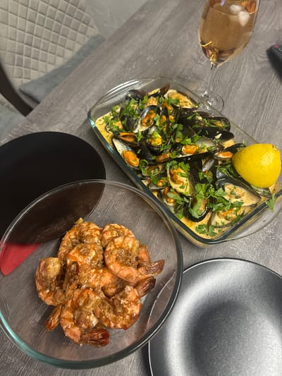 A glass baking dish filled with mussels in a creamy sauce, garnished with chopped parsley and a lemon wedge, is placed on a gray wooden table. To the left of the dish, a clear glass bowl contains cooked shrimp. A partially visible black plate and a wine glass with a light-colored beverage and ice are also on the table. The scene is set in a dining area with a quilted chair visible in the background. There are no people or animals in the image. The image does not provide information about the location in Sustaš, Montenegro.