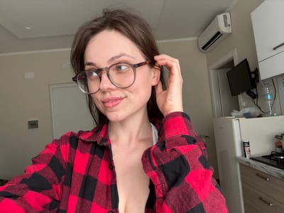 A female subject, appearing to be in her twenties, is depicted in an indoor residential setting in Scafati, Italy. She has brown hair and is wearing round, dark-rimmed prescription glasses. Her right hand is positioned near her right ear and the temple of her glasses. She is clad in an unbuttoned red and black plaid flannel shirt over a white undergarment. Her gaze is directed slightly to her right. The background features a cream-colored wall and a white ceiling. Visible objects include a white wall-mounted air conditioning unit on the upper right, and below it, a dark-colored flat-screen television attached to the wall with a mounting bracket. To the right of the subject, a white refrigerator stands next to a kitchen countertop with light-colored marbling. Above the countertop are white overhead cabinets, and on the countertop, a gas stovetop with black grates is partially visible. A clear plastic water bottle is present on the countertop, along with other small, indistinct items. A white door frame is visible in the middle ground, indicating an adjoining room.
