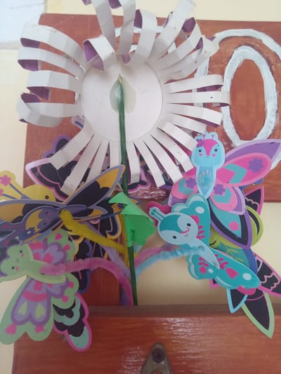 The image displays a close-up of a colorful craft project featuring several paper butterflies and a white paper flower with purple accents. The butterflies are made of layered cardstock with intricate designs and bright colors, including pink, purple, teal, and green. Some butterflies have googly eyes and antennae made of pipe cleaners. The flower appears to be a 3D construction with white petals and a green stem. The entire arrangement is attached to a wooden surface, which has an oval shape painted on it in brown and white. The background is a pale yellow wall. The specific location in Terebovlia, Ukraine, is not discernible from the provided image.