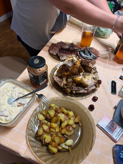 This media file captures a close-up overhead view of a table laid out with food and drinks. The setting appears to be a casual meal, likely indoors given the lighting. On the table, there are several dishes, including a whole roasted chicken, a plate of fried potatoes, a small bowl of fish, and a salad in a glass container. A jar of what appears to be a condiment is also visible. To the left, a person's arms are visible, holding a bottle of amber-colored liquid, presumably alcohol. A glass filled with the same liquid is also present. In the background, a pack of Sobranie cigarettes and a lighter are on the table, along with a mobile phone showing the time as 2:31. The text visible on the cigarette pack is in Ukrainian, warning about the dangers of smoking. The overall mood seems relaxed and convivial, suggesting a shared meal or gathering. The lighting suggests it might be evening or a dimly lit indoor space. The presence of a food spread with roasted chicken and potatoes indicates a hearty meal.