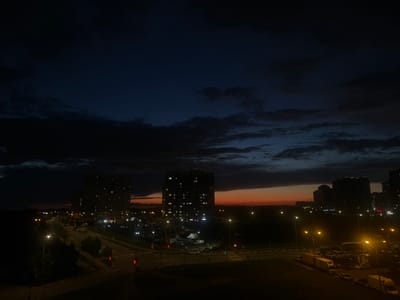 This is a nighttime photograph taken from an elevated vantage point, likely a building, overlooking a city scene in Domodedovo, Russia. The sky is a deep, dark blue, transitioning to hues of orange and red along the horizon, indicating either dawn or dusk. Dark, textured clouds are present in the upper portion of the sky.
In the mid-ground, several tall, multi-story apartment buildings are visible, their windows illuminated with warm light, suggesting occupancy. These buildings are silhouetted against the twilight sky. A road runs through the scene, lined with streetlights that cast pools of yellow light. Vehicles, including cars and vans, are present on the road and in parking areas. Some traffic lights are visible, displaying red and green signals.
The foreground shows a darker area, possibly a street or pavement, with shadows cast by the streetlights. The overall impression is of an urban environment at night, with the city lights contrasting against the darkening sky and the silhouettes of buildings. There are no identifiable people in the frame.