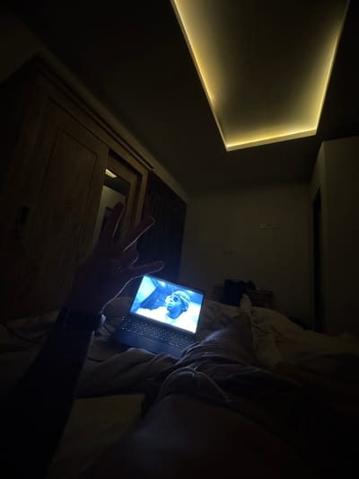 A low-angle interior shot shows a person lying in bed in Munggu, Indonesia. The room is dimly lit, with a recessed rectangular light source on the left wall. In the foreground, a hand is raised, displaying the "west side" hand gesture. A laptop rests on the bed, its screen illuminating the scene with a blue light. The screen displays a man wearing sunglasses and what appears to be a blue jacket. The person's arm, wearing a wristwatch, is visible in the lower right corner, alongside some light-colored bedding. In the background, there appears to be a wooden door or closet. Electrical outlets are visible on the wall.