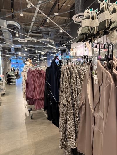 The image depicts the interior of a Primark retail store, specifically located in Philadelphia, United States. The scene is dominated by multiple clothing racks in the foreground and midground. On the right, a rack displays light mauve satin-like pajama sets, followed by a rack with leopard-print loungewear, and then dark blue satin-like pajama sets with white piping. Directly above these racks, bundles of ribbed socks in green and off-white are suspended from a circular fixture, bearing "Primark" branding. In the midground, a mobile clothing rack with wheels displays red and white striped pajama sets. Several partial mannequins are visible behind these racks; one wears a light purple patterned dress. Further into the store, additional clothing racks and shelving units displaying folded apparel are present on the left. The ceiling features an exposed industrial aesthetic with silver cylindrical ductwork, electrical conduit, and numerous track lighting fixtures. The flooring is a light gray tiled surface. In the distant background, a large window or glass facade reveals a blurred exterior urban environment or adjacent building, indicative of a city setting. No live individuals are visible.