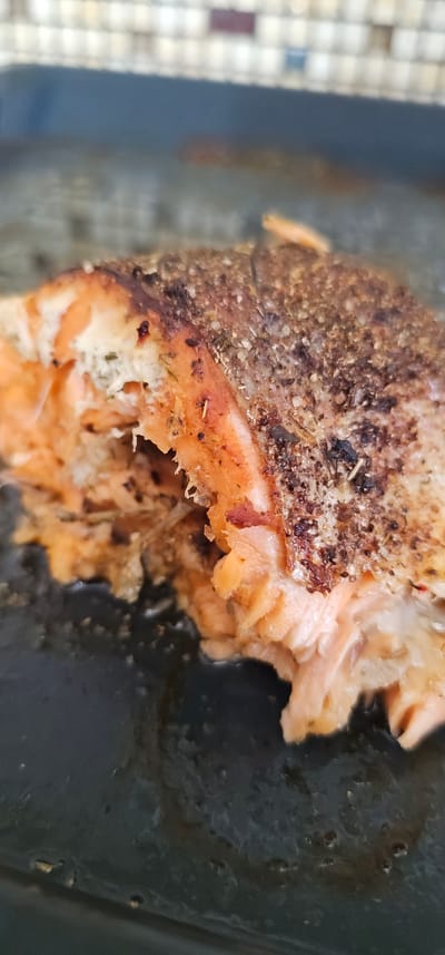 The image shows a close-up of a cooked piece of salmon. The salmon is seasoned with herbs and spices, giving it a browned and textured surface. The flesh of the salmon is a vibrant orange color and appears flaky. The salmon is placed in a dark cooking pan, which shows some residual oil and cooking residue. The background is blurred, but suggests a light-colored, tiled surface on the left and a dark, possibly metallic surface on the right. The lighting appears to be natural, coming from above and slightly to the left, casting shadows on the salmon. There is no text visible in the image. The setting appears to be a kitchen or dining area.