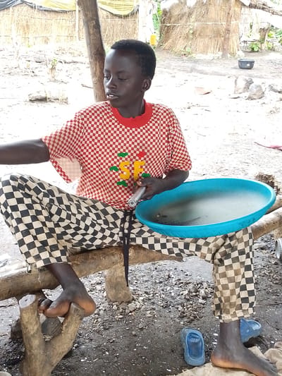 In the city of Otulu, Nigeria, a young individual is seated on a wooden bench. They are wearing a red and white checkered shirt with a red collar and checkered trousers in black and white. The individual is holding a metal rod that extends into a large, light blue circular basin filled with water. The basin rests on the bench next to the individual. The background shows a rustic dwelling constructed with natural materials, likely straw and wood, suggesting a rural or traditional setting. The ground is composed of dirt and small stones. A pair of blue flip-flops is visible near the bench. The overall scene depicts a moment of daily activity in a modest Nigerian environment.