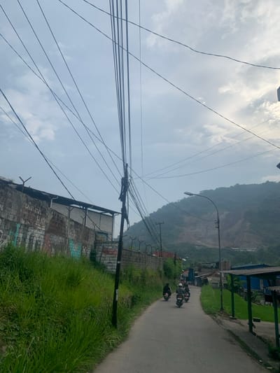 A street in Lagadar, Indonesia (latitude: -6.924, longitude: 107.538) is lined with a concrete wall on the left. The wall is covered in graffiti and topped with a barbed wire fence. Green grass grows up the hillside next to the wall. There are numerous utility wires running overhead. A group of people are riding motorcycles down the street. The street is paved and curves to the right, leading up a steep hillside. The hillside is covered in vegetation and a small rocky outcropping. A street lamp is mounted to a pole and casts a shadow on the street. The sky is overcast.