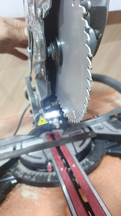 A close-up, downward-angled shot shows a miter saw with its blade prominently featured. The saw's metallic gray blade, with its sharp teeth, is the primary focus, positioned slightly off-center to the right. To the left of the blade, a clear plastic guard is visible, revealing a silver bolt and washer mechanism. Below the blade, a bright white light emanates from a small bulb, illuminating a yellow triangular safety symbol with a laser pointer beam. A dark grey power cord is coiled to the left of the saw. In the background, a blurred hand with visible fingers is seen reaching towards the saw. The saw is situated on a wooden surface, possibly a workbench, with fine sawdust scattered across it. The background is out of focus, showing hints of a wooden wall. The scene is devoid of any identifiable landmarks indicating a specific location within Pavino, Russia, or any other city.