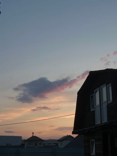 The image depicts a sunset scene in Solontsy, Russia. In the foreground, a dark, silhouetted wooden building with white-framed windows is partially visible on the right. To the left and in the background, several other structures, including houses with peaked roofs and what appear to be industrial buildings, are visible. A red wire stretches horizontally across the middle of the frame. The sky above is a gradient of pale blue and soft pink hues, with scattered clouds tinged with pink and orange, indicative of sunset. A dark, elongated cloud formation is prominent in the upper left quadrant. The overall impression is a rural or suburban landscape at dusk.