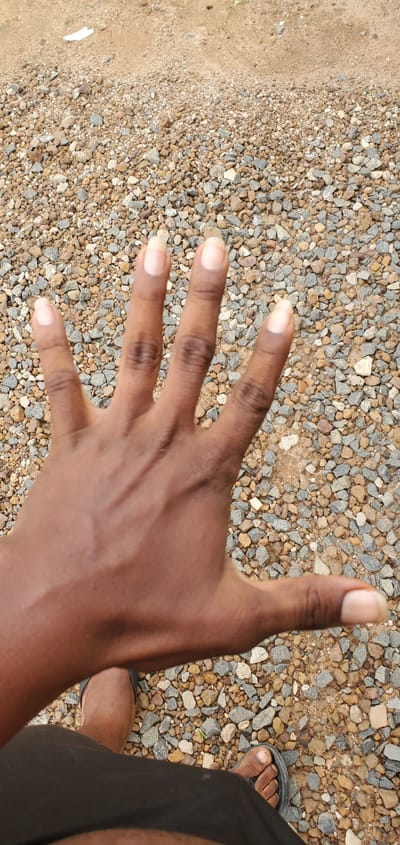 A person's hand, palm facing upwards and fingers spread, is shown in the foreground against a background of gravel and dirt. The hand is dark-skinned with lightly colored fingernails. The person is wearing black shorts and dark sandals. The scene appears to be outdoors, possibly in Numan, Nigeria, given the context.