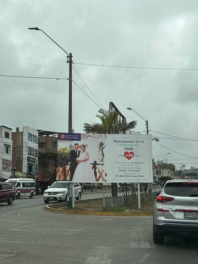 The image shows a street scene in Chorrillos, Peru, under a cloudy sky. In the foreground, a silver SUV is parked on the right side of the road, displaying a Peruvian license plate "BJY-413". Further down the road, a white minivan with red and blue stripes is visible, along with other vehicles and buildings. Dominating the center of the image is a large billboard advertising "Matrimonio Civil Comunitario 2026" (Community Civil Marriage 2026) sponsored by the "Municipalidad de Chorrillos" (Municipality of Chorrillos). The billboard features a photograph of a wedding couple and details about the event, including the date, time, and cost. A street lamp and power lines are also present in the scene.