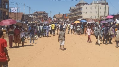 A large crowd fills a wide, dirt road in a town or village. Buildings are visible in the background. Many people are walking in the same direction, some carrying umbrellas for shade. The sky is clear and the lighting indicates it is daytime. One individual walks away from the viewer, down the center of the road.