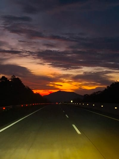 This is a photograph of a highway in Lamtamot, Indonesia, at dusk. The sky is filled with dramatic clouds painted in shades of orange, yellow, purple, and dark gray, indicating the sunset. The road, a dark asphalt surface, stretches into the distance, marked by a white line on the left and dashed white lines on the right. The headlights of the vehicle from which the photo was taken cast a bright yellow glow on the road in the foreground. Trees and hills are silhouetted against the colorful sky on either side of the highway. Small orange lights are visible along the side of the road, and further down the highway, there are intermittent yellow lights. A mountain range is visible in the background, partially obscured by the fading light.