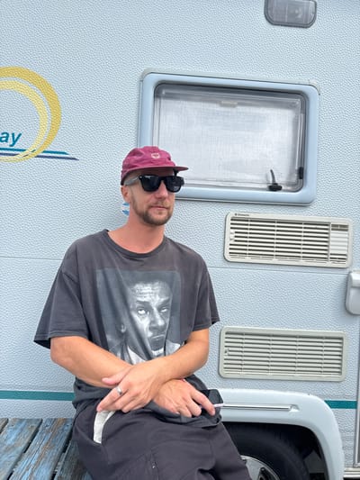 An image captured in Vessigebro, Sweden, shows a man posing relaxed against the side of a light blue-grey camper or caravan. The man, with a beard and wearing sunglasses, looks towards the camera. He is dressed in a maroon baseball cap and a dark grey t-shirt featuring a monochrome graphic of a person's face, which resembles Tupac Shakur. His arms are crossed over his chest, and he holds a smartphone in his right hand, revealing a silver ring on his left ring finger.
The camper's exterior has a distinct pebbled texture. Visible features on the camper include a rectangular window with a light grey frame and two vertically oriented white vents, each clearly displaying the brand name "Dometic". A white wheel arch covers part of a tire on the right side of the camper. Towards the bottom left, a yellow and blue graphic design with the partial word "ay" is visible on the camper's wall, suggesting part of a larger word like "Holiday" or "Sunny Day." At the bottom right of the frame, a weathered blue wooden structure, possibly a bench or step, is partially visible. The scene is set outdoors during daylight hours.