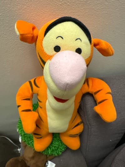 This is a close-up, vertical, eye-level shot of a plush toy Tigger character. The toy is predominantly orange with black stripes and a cream-colored belly. It has a round, yellow face with black eyes and a simple curved black line for a mouth. Its nose is a soft pink, fuzzy sphere. The toy is lying on its back, with its legs and arms splayed out. One of its front paws is visible on the left side of the frame, near the bottom. The toy is positioned on a gray, textured surface, possibly upholstery, with a hint of a lighter textured surface on the far left, which could be a wall. A small patch of bright green material, resembling grass, is visible under the toy's body. A brown plush toy can be seen partially in the background to the right. There is no discernible text in the image. The lighting appears to be ambient, with no strong shadows, suggesting an indoor setting. The overall impression is of a well-loved children's toy.