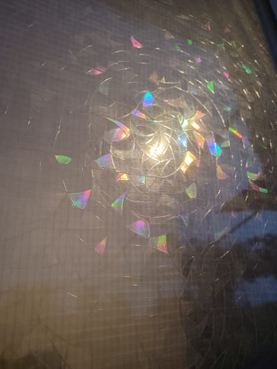 This image depicts a close-up view of a window with textured film, possibly a privacy or decorative film, applied to the glass. The film features a repeating pattern of intersecting lines that create geometric shapes, resembling a shattered glass effect or a stylized web. Light is passing through the film, causing a prismatic effect and casting colorful refractions, predominantly in hues of pink, blue, green, and yellow, onto the surface. The texture of the film itself is visible as a fine grid or weave beneath the larger pattern. The overall lighting suggests it is either early morning or late evening, with a strong light source coming from behind the window, illuminating the refractions. The environment outside the window is not clearly visible, appearing as a dark, blurred mass. This image was taken in Novosamarka, Ukraine.