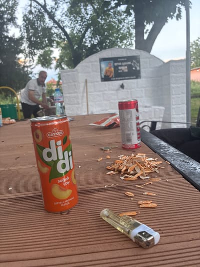 In Radievo, Bulgaria, on a wooden table, a can of "didi" peach-flavored iced tea by Çaykur stands prominently. Beside it, a red and white can of an unspecified beverage, possibly an energy drink, is present. A pile of what appears to be dried sunflower seed shells, along with loose shells and a few coins, are scattered on the table. A clear plastic lighter lies in the foreground. In the background, a man in a light-colored polo shirt and dark pants is partially visible, attending to something on the table. A bottle of Devin mineral water is also visible behind him. Further in the background, a white brick structure with a dark plaque bearing a photograph and text is mounted on a wall, with a tree providing shade. The scene appears to be an outdoor setting, possibly a park or a garden area.