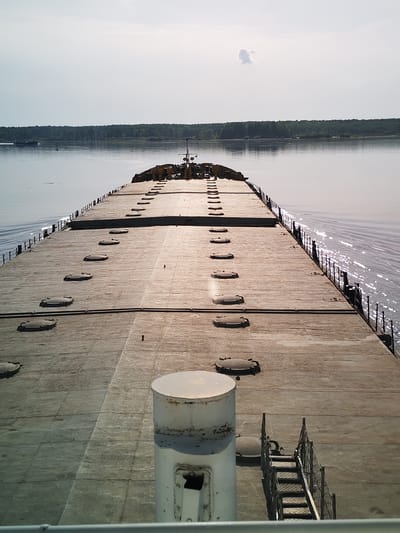 The image captures a wide-angle perspective from the deck of a large barge on the Sheksna River in Shelomovo, Russia. The barge stretches horizontally across the frame, its concrete surface marked with regularly spaced, round access hatches. A white cylindrical structure and a set of metal stairs are visible in the foreground on the right. The river, reflecting sunlight, extends to the horizon where a treeline is visible. A second, smaller vessel appears in the distance on the left. The sky is overcast with a single small cloud.