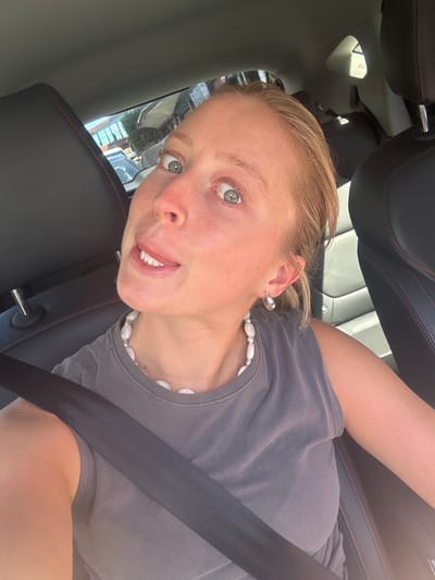 A fair-skinned adult female is depicted in a selfie taken from the front passenger seat of a vehicle during daylight hours. She is looking directly at the camera with wide blue-grey eyes and her tongue slightly extended between her lips. Her light-colored hair is pulled back. She wears a dark grey sleeveless top, a necklace composed of irregular white beads, and silver-toned hoop earrings. A dark grey seatbelt diagonally crosses her torso and shoulder. The car's interior features black seats with red stitching on the headrest and seat edges, along with a textured, ribbed seat back. Bright sunlight is visible from the right side of the frame, illuminating the subject. Through the vehicle's rear window, blurred residential buildings with pitched roofs, trees, and a partially visible parked white vehicle are discernible, indicative of an urban or suburban street. The specific location within Wollongong, Australia, is not identifiable from the visual evidence provided.