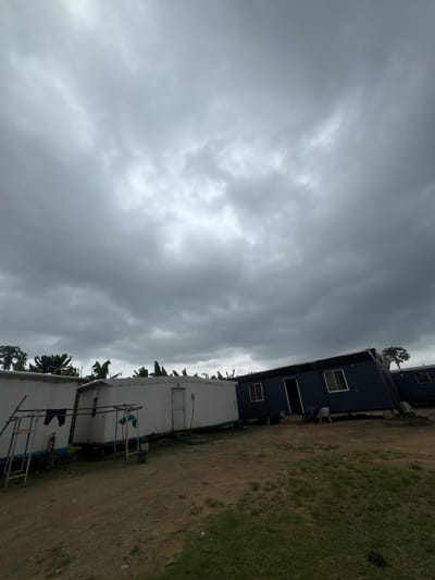 The image shows temporary buildings at a site in Igwuruta, Nigeria under a cloudy sky. The buildings are primarily single-story structures, with a mix of dark and light-colored exteriors. The surrounding ground is a combination of dirt and grass. There is a person visible entering one of the buildings. Laundry is hanging outside one of the light-colored buildings. There is a fire extinguisher next to the light-colored building.