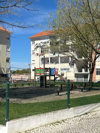 The image depicts a residential area in Seixal, Portugal, characterized by apartment buildings with clotheslines stretched between balconies. The buildings are light-colored with red-tiled roofs. A wire fence separates the buildings from a green space and a paved sidewalk. A tree with bare branches is visible in the foreground. The sky is blue with some wisps of clouds. No people are clearly visible in the image.