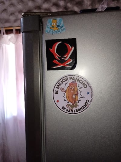 The image shows a close-up of a silver refrigerator door in a home in El Talar, Argentina, located at latitude -34.473 and longitude -58.639. The door has three stickers. The top sticker features a cartoon hot dog in a red cap and a yellow shirt, labeled "El Mejor Pancho de San Fernando." The sticker below it features a red and black stylized "X" in a black circle. The lowest sticker shows a cartoon depiction of Bart Simpson, with the word "Relax" in a bubble above him. The refrigerator door is slightly ajar. The background is out of focus and blurry, revealing a white wall. The refrigerator is likely part of the kitchen, and the scene suggests the presence of a person who has decorated the appliance.