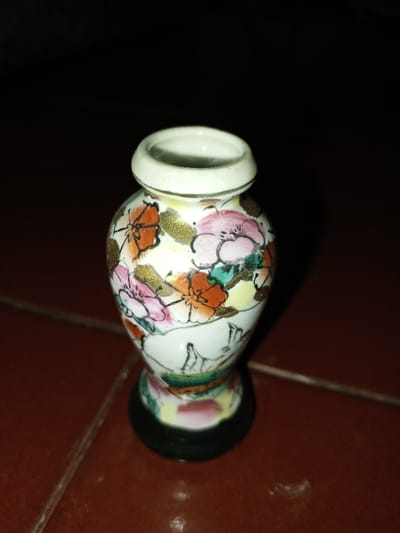 A miniature, white ceramic vase rests on a reddish-brown tiled surface. The vase features a globular body, a narrow neck, and a flared rim, supported by a distinct black circular base. Its surface is adorned with hand-painted floral motifs in various colors, including prominent pink and orange blossoms, along with teal-green leaves and yellow accents, all outlined in dark pigment and embellished with gold-colored details. The background is uniformly dark and out of focus. No other subjects, actions, or interactions are visible. The specific location in the city of Longmiang, Laos, is not discernible from the visual information provided.