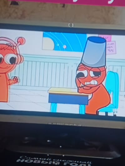The image depicts a screenshot from a cartoon, displayed on a flat-screen television. Two cartoon characters are visible. On the left is an orange, anthropomorphic character with large eyes, antennae, and headphones. This character appears to be looking towards the right. On the right, another orange character is seated at a desk with a blue chair. This character is wearing a grey bucket on its head and appears to be in distress, with its hands near its face and its mouth open in a grimace. The background is a pale blue wall with vertical lines and some abstract shapes. The television screen is the primary focus, and the bottom portion of the image shows the dark surface of the television stand, with some text visible in a silvery font, reading "САМЫЙ СЧАСТЛИВЫЙ НОВЫЙ ГОД!" which translates to "HAPPIEST NEW YEAR!". The location in Berezivka, Ukraine, is not discernible from the image content.