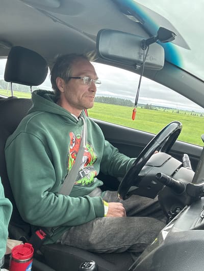 A Caucasian male with short, light brown hair and glasses is driving a vehicle. He is wearing a green hoodie with a graphic print and a grey seatbelt. The interior of the car is visible, including the steering wheel, rearview mirror, and dashboard. Outside the car, a green, rural landscape with trees and fields is visible through the windshield and side window. The sky is overcast. A red insulated cup and part of another person's arm are visible in the foreground.