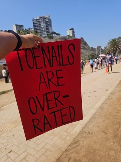 A person stands holding a red sign that reads "TOENAILS ARE OVER-RATED". The person is wearing a black wristband. The background suggests the city of Chorrillos, Peru, with a palm tree, buildings, and a slight cliff face visible in the background. There is a group of people standing in the distance. The ground is made of light-colored brick.