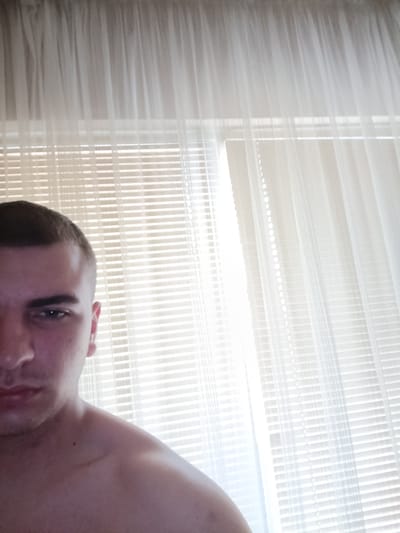 This partial indoor portrait, taken in Rakovski, Bulgaria, features a young man occupying the left side of the frame. Only his left eye, part of his nose, mouth, and chin are visible, giving him a neutral or slightly serious expression. He has a short, dark buzz cut. His bare left shoulder and upper chest are also in view, revealing a faint, short scar near his collarbone. The background is a brightly illuminated window covered by horizontal white venetian blinds and soft, translucent sheer curtains. Strong daylight streams through these coverings, suggesting it is daytime. The man is posing for the camera; no other specific activity is depicted. There is no visible text in the image.