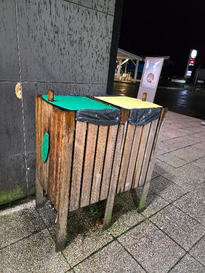 The image captures an outdoor scene at night in Longeville-lès-Saint-Avold, France, depicting a weathered wooden slatted bench. The ground is paved with speckled, light-colored tiles that appear wet, suggesting recent rain or damp conditions.
The bench is the central subject, with two black plastic bags draped over its back and seat, covering parts of it. Beneath these bags on the left side of the bench, a bright yellow panel and a green panel are visible, possibly forming a makeshift structure or cover. A small, oval green shape is affixed to the visible side leg of the bench.
To the left of the bench is a dark, textured wall or building. In the background, beyond this wall, artificial lights illuminate a commercial establishment, possibly a gas station or shop, with a prominent white and red illuminated sign that includes indistinct letters like "T" and "0". A white rectangular signboard with unreadable text is also visible in the distance.
The scene is devoid of people. The overall impression is of an outdoor public or commercial area late at night, under wet weather conditions.