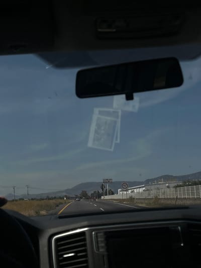 The image depicts a roadside scene from the interior perspective of a vehicle traveling on a multi-lane highway in Abasolo, Mexico. The top portion of the vehicle's dark interior roof liner and the rearview mirror are visible. Two rectangular, light-colored objects, possibly photographs, are adhered to the windshield below the rearview mirror. The driver's side of the steering wheel and a portion of the dashboard, including air vents, are present in the lower frame.

The highway ahead extends into the distance, featuring a yellow dividing line and dry, vegetated shoulders. On the left side, utility poles and power lines run parallel to the road. Several vehicles are observed traveling on the highway in the same direction. On the right, a white circular speed limit sign indicating "40" is visible. Behind this sign, a large, light-colored commercial or industrial building with a security fence is present, along with a larger sign structure. In the background, a range of mountains or hills is visible under a blue sky with scattered light cloud formations. No individuals are clearly identifiable, and the primary action is vehicular travel.