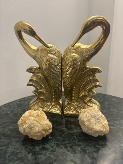 This media file features a brass swan figurine with two rough, light-colored stones. The swan is highly detailed, with visible feathering and an ornate crest. The figurine is positioned on a reflective surface that creates a mirrored image of the swan and the stones. The background consists of a white wall and a dark green, marbled surface, likely a tabletop. The lighting suggests an indoor setting, possibly during the daytime with artificial light contributing. The stones appear to be natural, unpolished, and rough in texture, with visible crystalline structures. The brass swan has a lustrous, polished finish, reflecting its surroundings. The overall scene is static, with no discernible activity other than the arrangement of the objects. The location is specified as Delray Beach, United States of America, but there are no specific visual cues in the image to confirm this. There is no visible text in the media file.